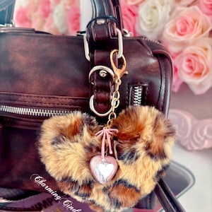 May include: A brown and tan leopard print heart-shaped keychain with a pink bow and a heart-shaped charm featuring a unicorn design. The keychain is attached to a brown handbag.