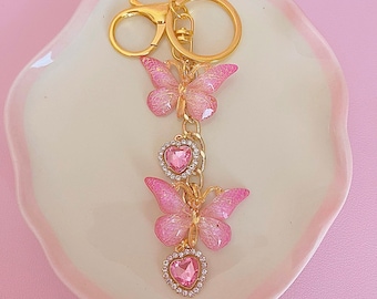 May include: A gold-colored keychain featuring two pink butterfly charms and two heart-shaped charms. The butterfly charms have glittery wings and are connected by a gold chain. The heart charms are encrusted with small, clear rhinestones. The keychain is on a white, heart-shaped dish.