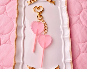May include: A pink heart-shaped lollipop keychain with a gold chain and a gold heart-shaped clasp. The keychain is displayed on a white tray with gold trim and two gold bows.