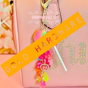 May include: A pink handbag with gold hardware and a charm. The charm features a heart-shaped pendant, gummy bear charms, and a lollipop. The bag has the word "Juicy" in rhinestones. The text "SHOPPINGPAGEXO" is also visible.