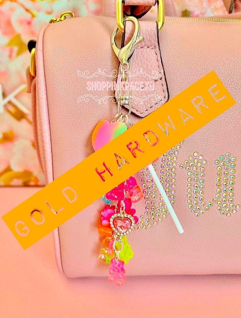 May include: A pink handbag with gold hardware and a charm. The charm features a heart-shaped pendant, gummy bear charms, and a lollipop. The bag has the word "Juicy" in rhinestones. The text "SHOPPINGPAGEXO" is also visible.