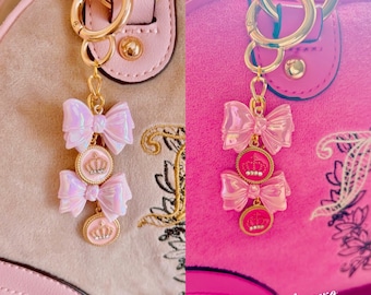 Può includere: Due borse a mano rosa con catene e ciondoli dorati. I ciondoli sono fiocchi rosa e bianchi con accenti dorati a forma di corona. Il testo '@shoppinkpagexo' è visibile nell'angolo in basso a destra dell'immagine.