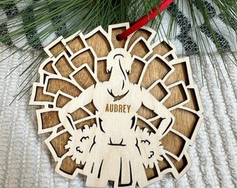 May include: Wooden Christmas ornament shaped like a cheerleader with pom-poms. The ornament is brown and has the name "Aubrey" engraved on it.