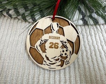May include: Wooden soccer ornament with a player silhouette holding a ball. The ornament is brown with a gold number 26 and the name "Beckham" on the player's jersey.