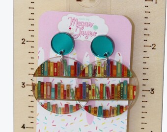 May include: Clear acrylic earrings with a bookshelf design, featuring a teal stud and a half-circle shape. The earrings are displayed on a wooden stand with the text "Made By Megan Jayne". The earrings are on a pink card with the text "Megan Jayne".