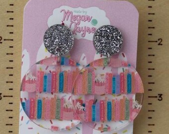 May include: Clear acrylic earrings with a silver glitter top and a colorful book design. The earrings are displayed on a wooden stand with the text "Made By Megan Jayne". The card has the text "Megan Jayne" and the website address.