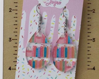 May include: A pair of clear, teardrop-shaped earrings featuring a colorful bookshelf design. The earrings are displayed on a pink card with the text "Megan Jayne" and a wooden display stand with the text "Made By Megan Jayne".