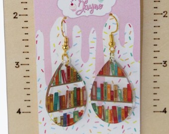 May include: Teardrop-shaped earrings with a bookshelf design, displayed on a wooden stand with a ruler. The earrings have gold-colored hooks and are on a pink backing with the text "Made by Megan Jayne". The stand has the text "Made By Megan Jayne".
