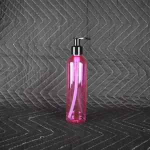 Puede incluir: Una botella de plástico rosa con una bomba de plástico negra. La botella está vacía y se encuentra sobre una superficie acolchada negra.
