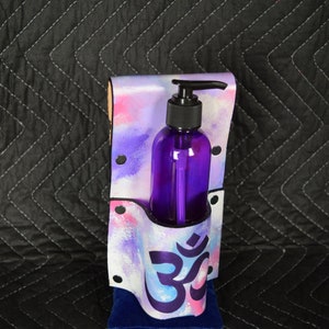 May include: A purple bottle with a pump dispenser is held in a white and pink holder with a blue and purple Om symbol. The holder is sitting on a blue velvet cushion.