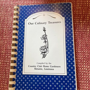Może przedstawiać: Niebieska i biała książka kucharska zatytułowana "Our Culinary Treasures" skompilowana przez Country Club Home Gardeners z Metairie w Luizjanie. Okładka przedstawia czarno-białą ilustrację kwiatu.