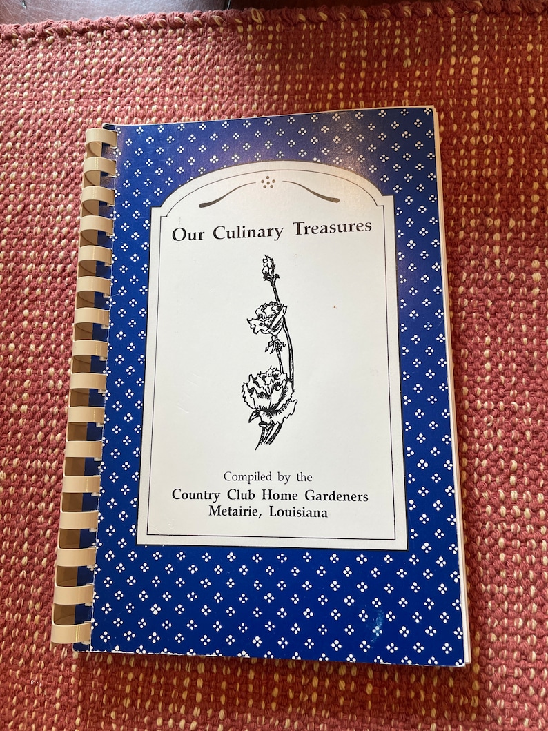 Może przedstawiać: Niebieska i biała książka kucharska zatytułowana "Our Culinary Treasures" skompilowana przez Country Club Home Gardeners z Metairie w Luizjanie. Okładka przedstawia czarno-białą ilustrację kwiatu.