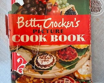 May include: A vintage red cookbook with the title "Betty Crocker's Picture Cook Book". The cover features a colorful still life of fruit and a tiered dessert stand with various pies and tarts.