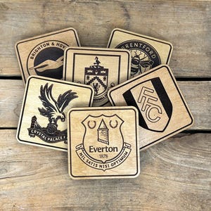 May include: A selection of wooden coasters displaying football club emblems. The coasters feature logos for Brighton & Hove, Brentford, Crystal Palace, Everton, and Fulham. Each coaster is square with a light wood grain finish.