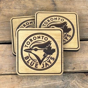 May include: Three square wooden coasters featuring the Toronto Blue Jays logo. The logo includes a blue jay bird and the words "Toronto Blue Jays" in a circular design. The coasters are made of light-coloured wood with a dark border.