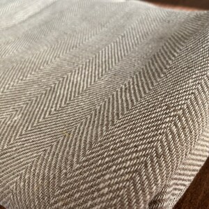May include: A close-up of a beige herringbone fabric with a subtle texture. The fabric is woven with a repeating pattern of diagonal lines.