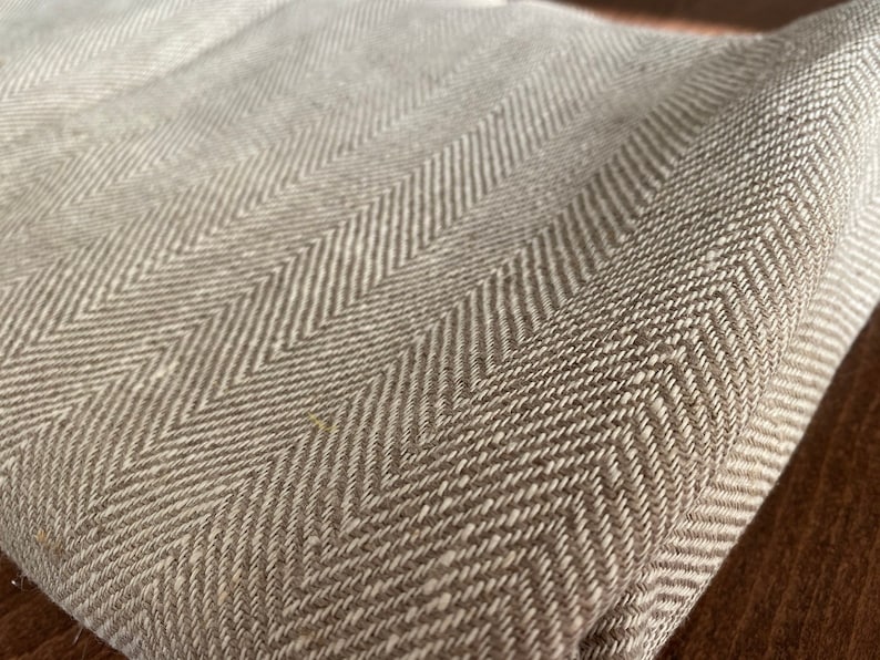 May include: A close-up of a beige herringbone fabric with a subtle texture. The fabric is woven with a repeating pattern of diagonal lines.