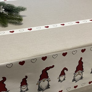 Può includere: Una tovaglia beige con un bordo bianco con cuori rossi e grigi. Il bordo inferiore mostra gnomi con cappelli rossi e barbe bianche. Un rametto di sempreverde e un ornamento a fiocco di neve argentato sono nell'angolo in alto a sinistra.