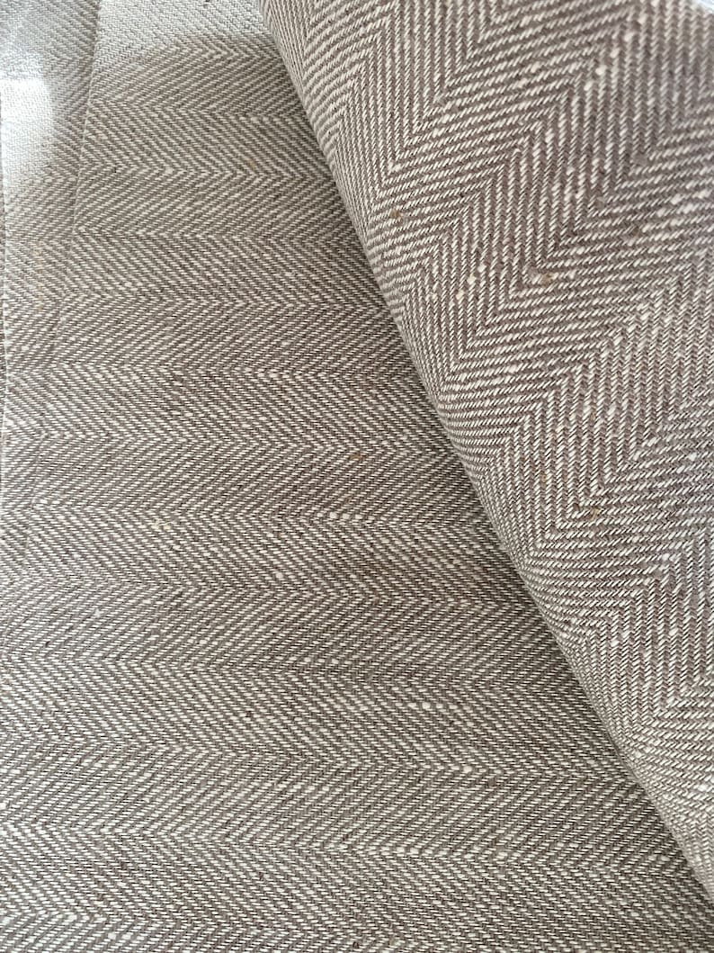 May include: Close-up of a brown and white herringbone fabric. The fabric is woven with a subtle texture and has a slightly rough feel.
