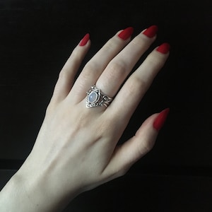 May include: A silver ring featuring a white oval stone and elaborate detailing. The ring is worn on a hand with red nail polish.