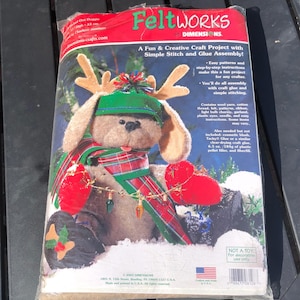 May include: A Feltworks craft kit featuring a plush dog dressed for the holidays. The dog wears a green hat, a red and green plaid scarf, and antlers. The kit includes materials for simple stitch and glue assembly.