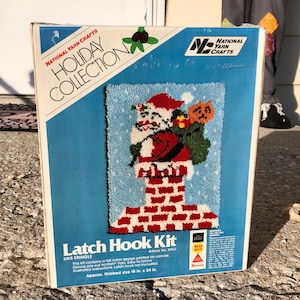 May include: A vintage National Yarn Crafts Holiday Collection latch hook kit. The kit features a design of Santa Claus in a chimney, with a finished size of 18 in. x 24 in. The box has a blue background with the text "Latch Hook Kit".