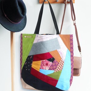 Könnte beinhalten: Eine Patchwork-Tragetasche mit verschiedenen Stoffen und Farben, darunter Rot, Blau, Grün und Gelb. Die Tasche hat einen schwarzen Henkel und einen braunen Riemen. Die Tasche hängt an einem Holzhaken.