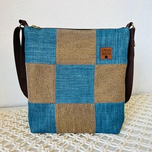 Könnte beinhalten: Quadratische Umhängetasche mit Patchwork-Design in Blau- und Beigetönen. Die Tasche hat einen braunen Schulterriemen und einen Reißverschluss. Ein kleines Lederetikett trägt die Aufschrift "LANA WERK".