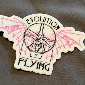 May include: A white sticker with a black and pink illustration of the evolution of flying. The illustration shows a propeller, wings, and the text "Evolution of Flying".