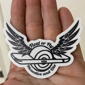 May include: A white sticker featuring a black graphic of a one-wheel electric skateboard design with wings. The text "Float or Fly" is above the wheel, and "Nosedive and Die" is below. The sticker is held in a hand.
