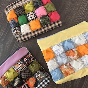 May include: Three square zippered pouches featuring a quilted puff design. Two pouches display a mix of Halloween-themed fabrics with black and white buffalo check borders. One pouch features a yellow border with light blue and floral fabric squares.