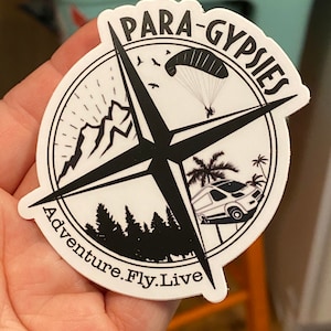 May include: A white round sticker with a black compass design. The compass points to "Adventure. Fly. Live." and has a paraglider, mountains, trees, and a van. The text "Para-Gypsies" is written around the compass.