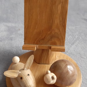 Peut inclure: Un support de téléphone en bois avec une figurine de lapin et de tortue sculptées. Le support est en bois brun clair et possède un support de téléphone rectangulaire.