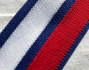 May include: A close-up of a white fabric with a blue and red striped ribbon sewn on it. The ribbon is diagonal and the colors are bright.