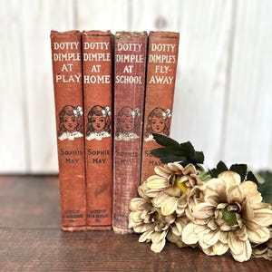 May include: Four vintage hardcover books titled Dotty Dimple at Play, At Home, At School, and Fly-Away by Sophie May, featuring reddish-brown spines with illustrations of a girl, accompanied by a cluster of cream-colored artificial flowers.