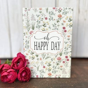May include: A rectangular sign with a cream-colored background featuring a colorful wildflower pattern in shades of pink, yellow, and green. Centered is a white label with a pink border containing the black text "oh HAPPY DAY".