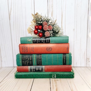 May include: A stack of five vintage hardback books with green and red spines, topped with a decorative cluster of artificial pine needles, gold leaves, and red berries. The titles include Crime and Punishment, Joan of Arc, and The Black Cauldron.