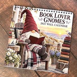 Può includere: Un calendario da parete 2027 intitolato Book Lover Gnomes che raffigura uno gnomo con cappello rosso e sciarpa a quadri che legge un libro su una sedia di legno affacciata sul mare al tramonto, con pile di libri, fiori e un gabbiano.