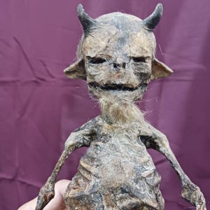 May include: A detailed, realistic sculpture of a small, skeletal creature with horns, a beard, and a weathered, aged appearance. The sculpture is made of a dark brown material and has a textured surface.