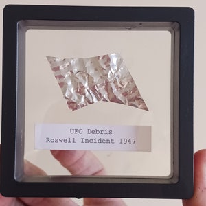 May include: A piece of silver-colored metallic debris in a black frame with a white label that reads "UFO Debris Roswell Incident 1947".