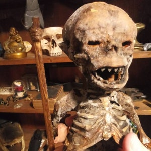 May include: A detailed, weathered skeleton figure with a spear. The figure has a skull-like head with visible teeth and eye sockets. The spear has a gray blade and a wooden shaft. The figure is held in a person's hand.