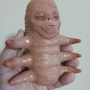 Pode incluir: Uma pequena figura de cerâmica bege de uma criatura com um rosto humanoide e quatro braços. A criatura tem os olhos fechados e uma expressão serena.