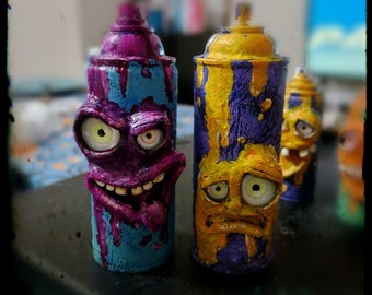 May include: Two spray paint cans with cartoon faces. The can on the left is blue, purple, and yellow with a wide, toothy grin and a purple tongue sticking out. The can on the right is purple, yellow, and blue with a sad expression and yellow eyes.