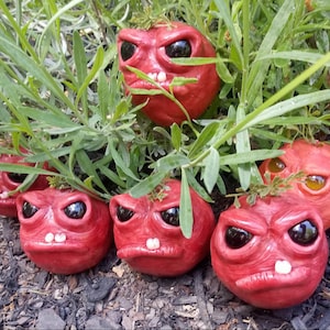 May include: A group of seven red, hand-painted, ceramic figurines with large eyes and toothy grins. The figurines are arranged in a cluster on a bed of brown mulch and green grass.