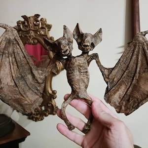 May include: A two-headed bat figurine with brown wings and a skeletal body. The figurine is held in a person's hand.