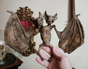 Può includere: Una statuetta di pipistrello a due teste con ali marroni e un corpo scheletrico. La statuetta è tenuta nella mano di una persona.