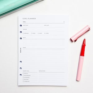 May include: A white goal planner with a black and grey gradient design. The planner has sections for goal setting, start date, goal date, reward, why, how, smart goal, and reflection. The planner has a dotted line design and a hole punch on the left side.