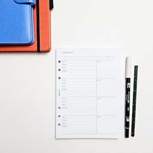 May include: A white page with the word "CONTACTS" at the top. The page is divided into four sections with columns for name, phone, email, address, and notes. There are two black markers and a white marker on a white surface.