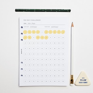 May include: A white 100-day challenge planner with a goal of 10K+ steps, a pencil, a triangular eraser, and a green ruler. The planner has yellow circles to track progress, with start and finish dates of 01/07/2025 and 30/07/2025.