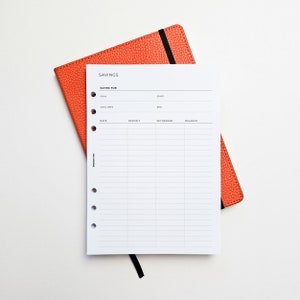 May include: A white printable savings tracker sheet with a black header that says "SAVINGS". The sheet has columns for date, deposit, withdraw, and balance. The sheet is lying on top of a bright orange leather-bound notebook.